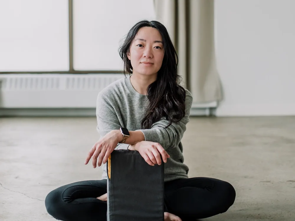 Caren Hui sitting cross-legged on the studio floor, arms resting on an upright bolster, looking into the camera.