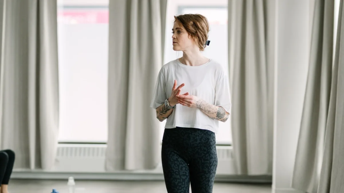 A teacher stands barefoot in the bright studio, hands together at her waist, looking off to one side as she speaks to the class.